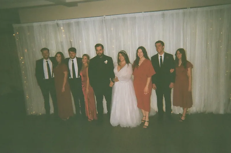 Nine people in formal wear stand in a row in front of a white curtain with string lights: men in dark suits and ties, and women in rust, orange, red and white dresses.