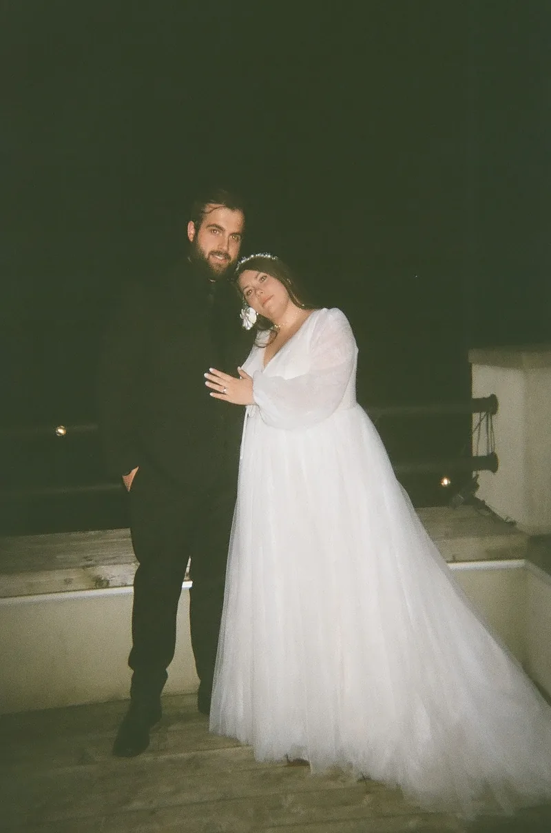 A bearded man in black and a woman in a white dress with a tiara and a flower pose close together on a terrace at night, with a dark sky behind them.