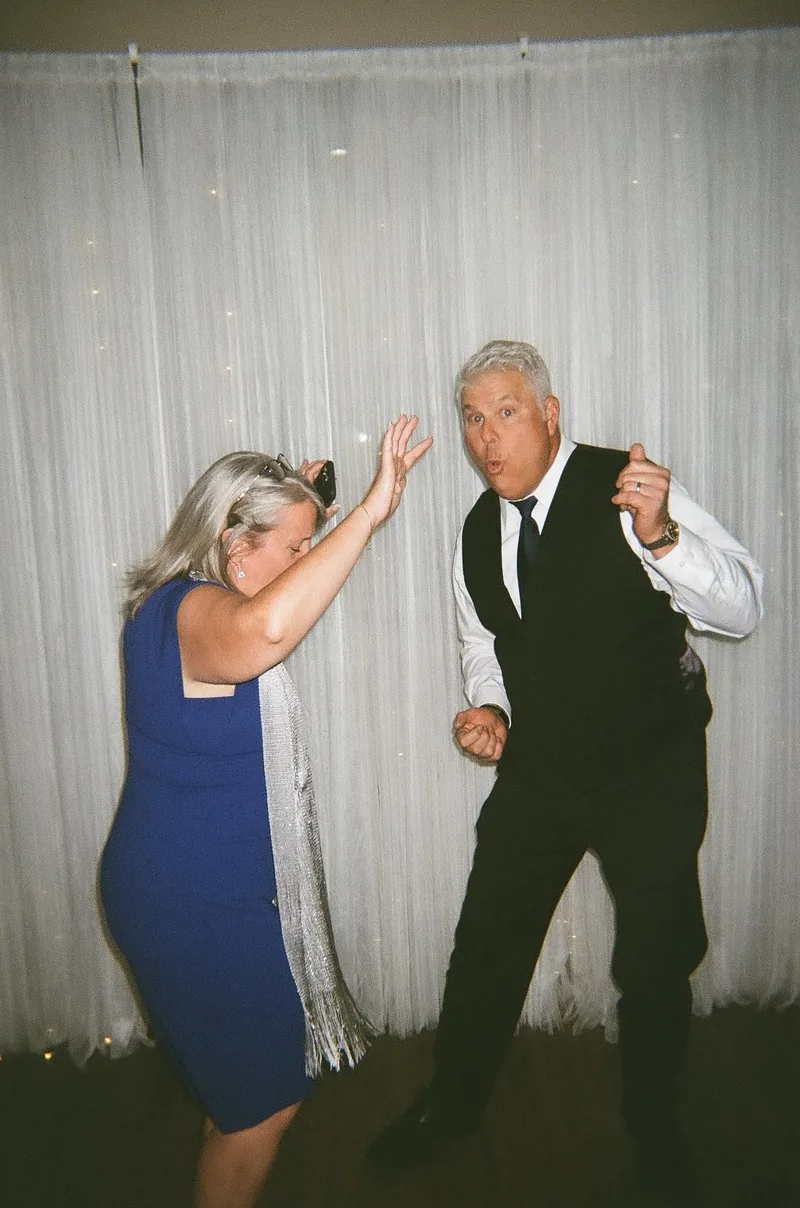 A woman in a blue dress and an older man in a vest and white shirt dance in front of a white curtain, both with an arm raised.