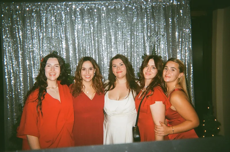Five women in red and white dresses stand close together smiling in front of a silver sequin curtain.