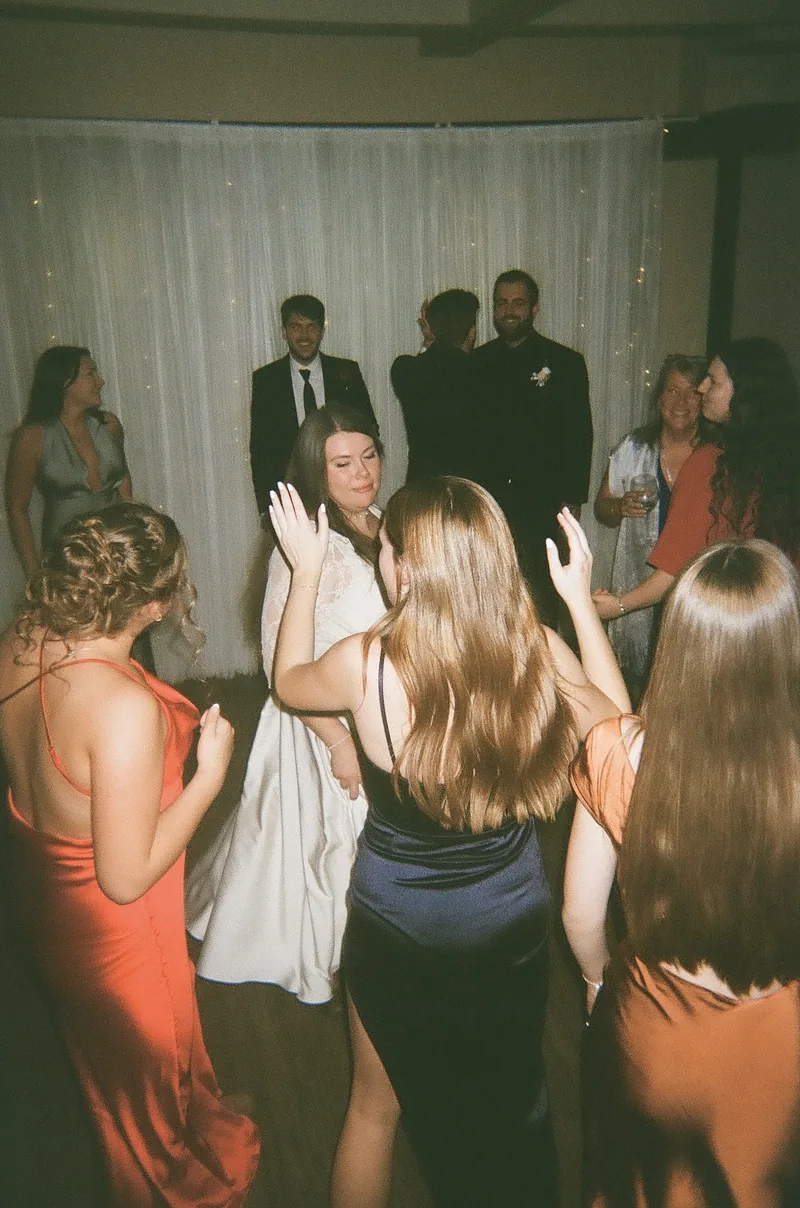 Guests dance on a floor in warm light, seen from behind, with women in red and navy dresses in the foreground and people in dark suits and a white dress standing near a white curtain at the back.