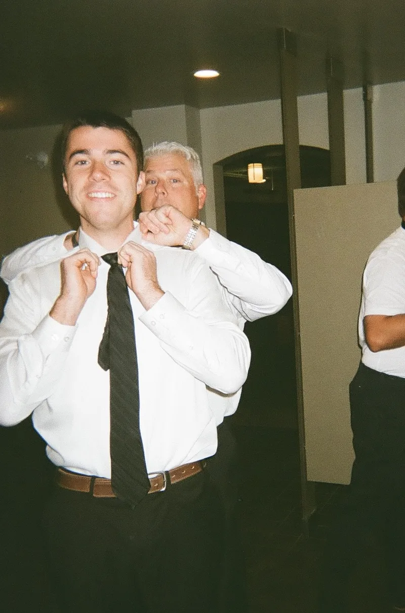 A smiling man in a white shirt and black tie adjusts his collar with both hands while an older man in a white shirt stands behind him, in a warm-lit room.